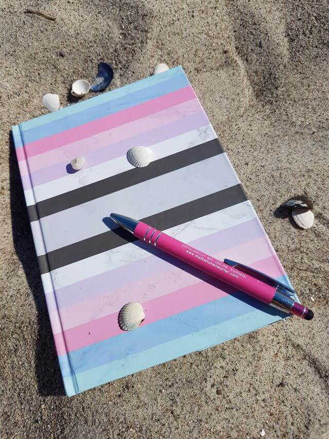 Ein gestreiftes Notizbuch liegt dekoriert mit Kugelschreiber und Muscheln am Strand